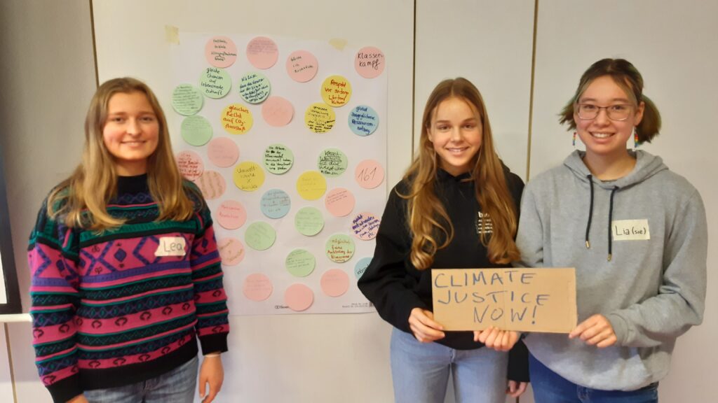 Wir (Lea, Paula, Lia) vor einer Wand mit angepinnten Zetteln. Wir halten ein Pappschild mit der Aufschrift Climate Justice now!